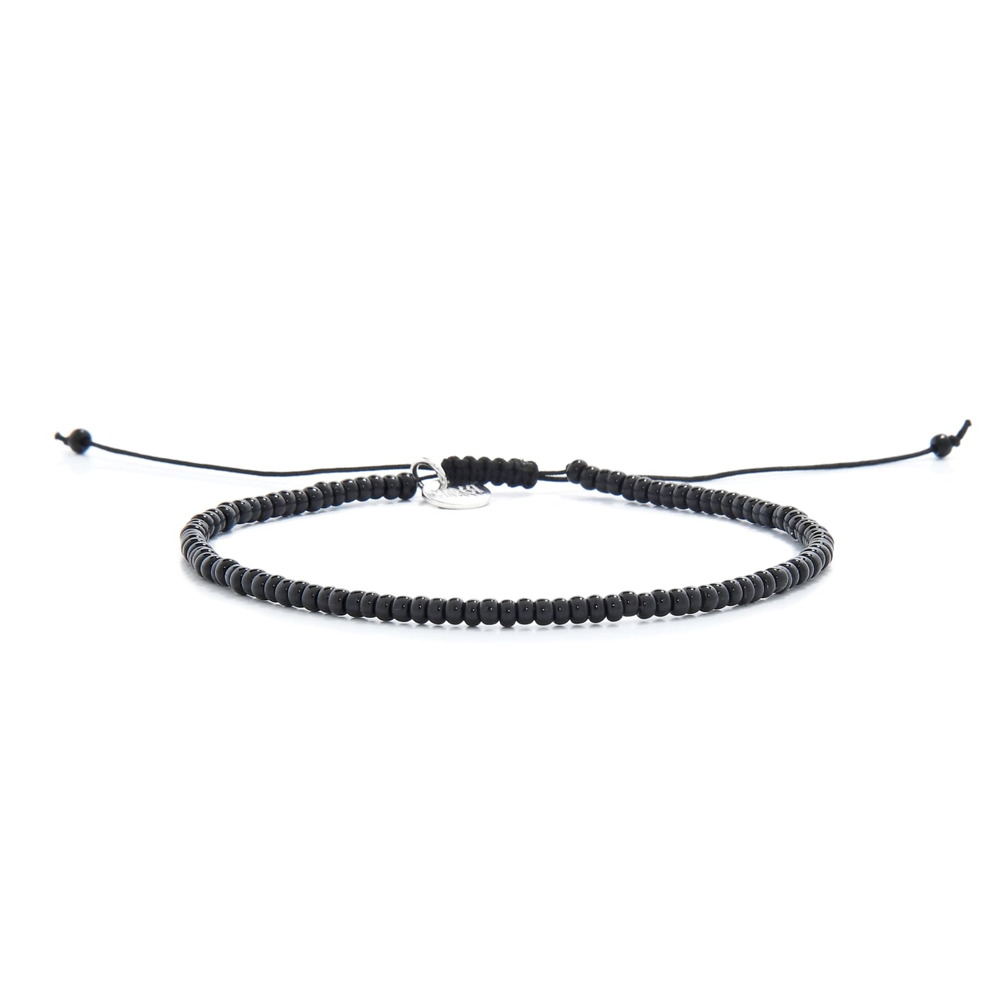 A full view of a simple black bead bracelet with an adjustable knot closure and a silver charm, lying on a white surface.