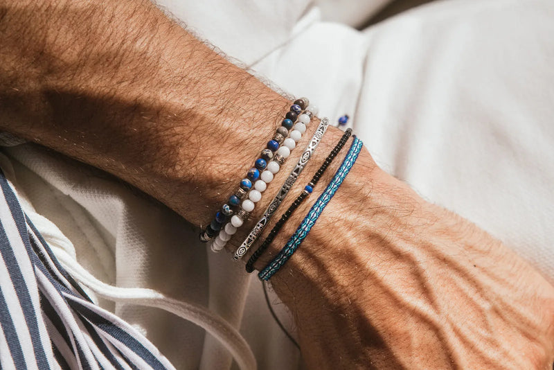 A silver cuff bracelet is layered with beaded and woven bracelets in a relaxed men’s summer stack.