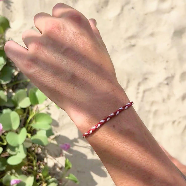 Hand wearing a red and white braided bracelet on a sandy background