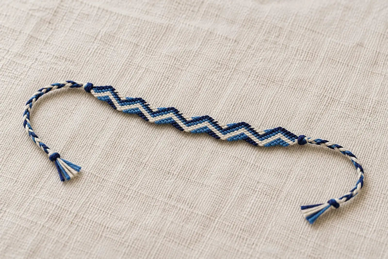 Zig Zag Friendship Bracelet: Finished Braided Ties Navy, ivory, and blue zig zag friendship bracelet laid flat on linen with braided ties and knotted ends.