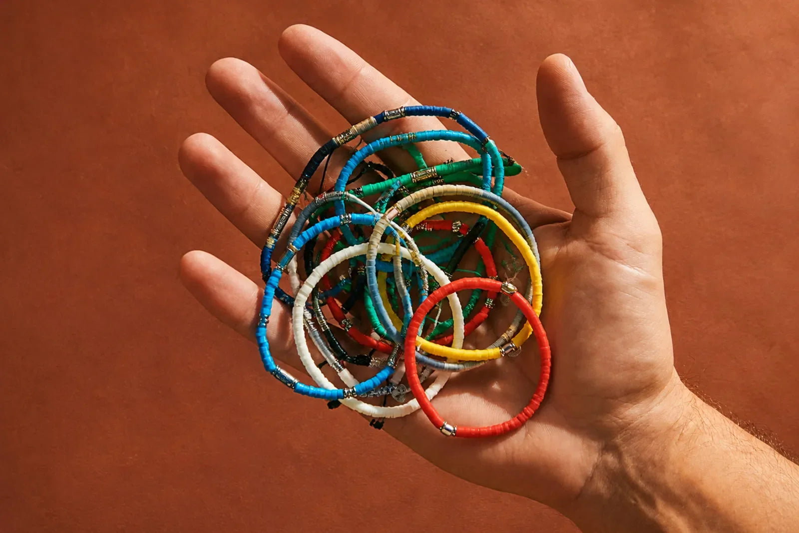 Clay bead bracelet collection in bright colors held in a man’s hand against a warm terracotta background.
