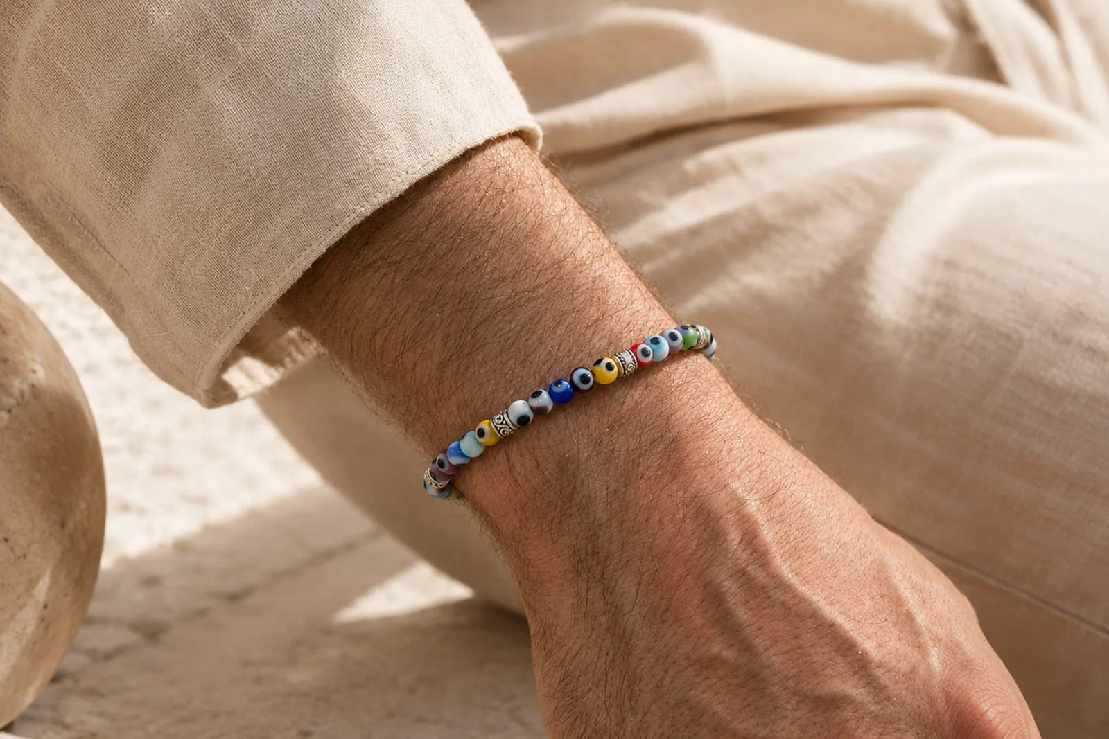 Evil eye bracelet on a man’s wrist, styled with beige linen in a warm Mediterranean setting.