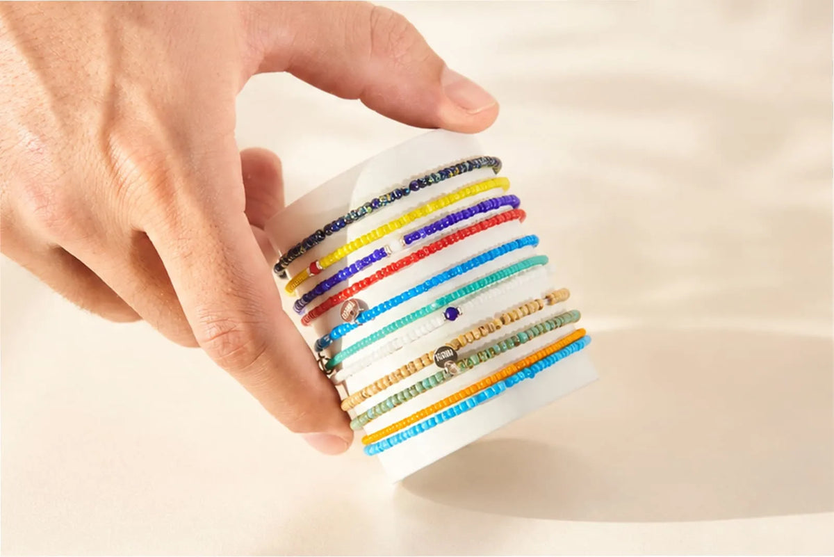 Glass Bead Bracelets displayed in colorful rows on a white card, held by a man’s hand against a neutral background.