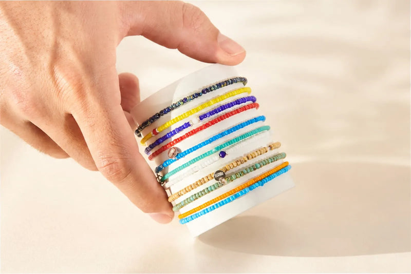 Glass Bead Bracelets displayed in colorful rows on a white card, held by a man’s hand against a neutral background.