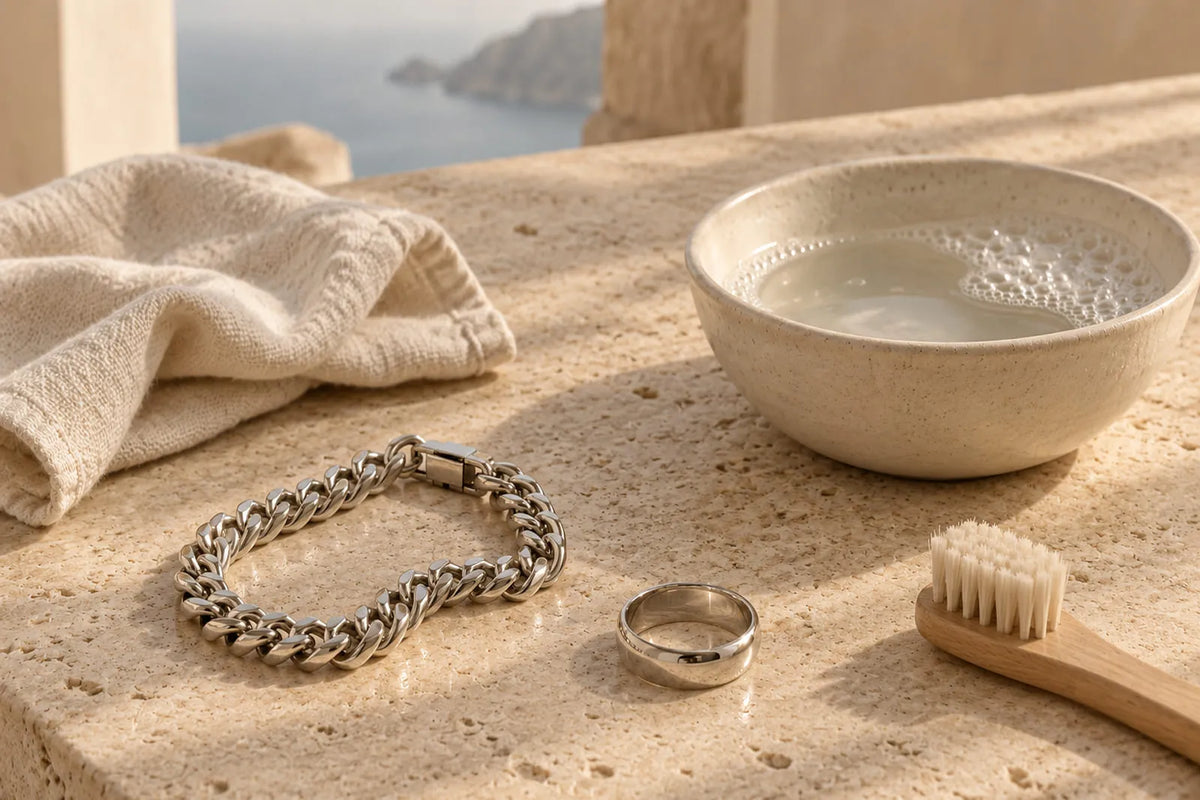 Silver chain bracelet and men's ring beside a bowl of homemade jewelry cleaner made with mild soapy water, a soft brush, and a drying cloth.