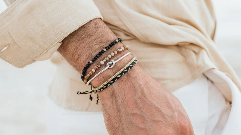 Men wearing a layered bracelet stack with neutral beads, silver accents, and a beige linen outfit