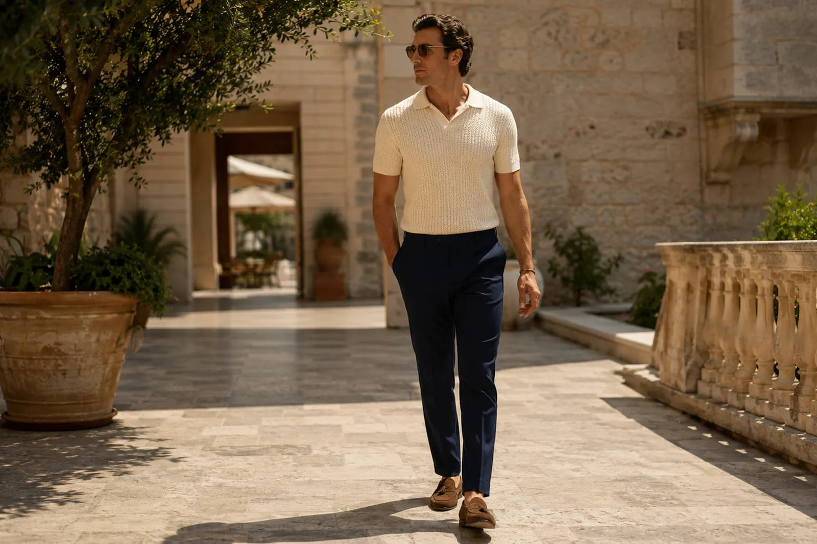 Man wearing a cream knit polo, navy tailored trousers and brown loafers in a Mediterranean courtyard, showing old money style for men.