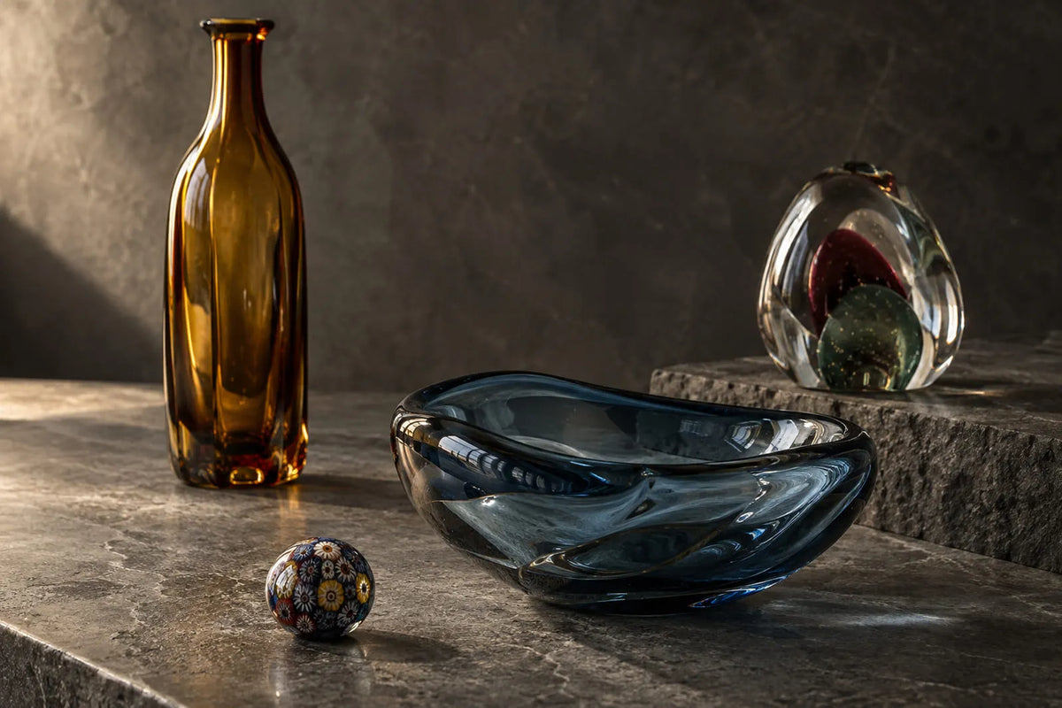 What Is Murano Glass illustrated with an amber bottle, smoky blue blown glass bowl, millefiori bead, and solid glass paperweight.