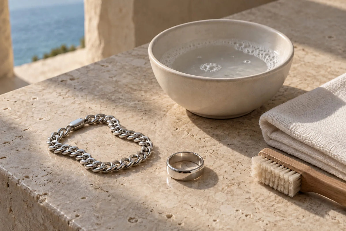Silver chain bracelet and ring beside a bowl of soapy water, a brush and a towel for cleaning silver jewelry.