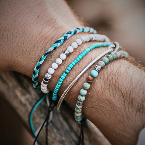 Close-up of a man's wrist wearing five stacked bracelets in turquoise, white, silver, and blue tones, featuring beads, cords, and a knot detail.