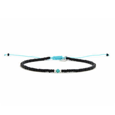 A straight-on view of an adjustable beaded bracelet with black beads, a turquoise bead, and a bright blue cord on a white surface.
