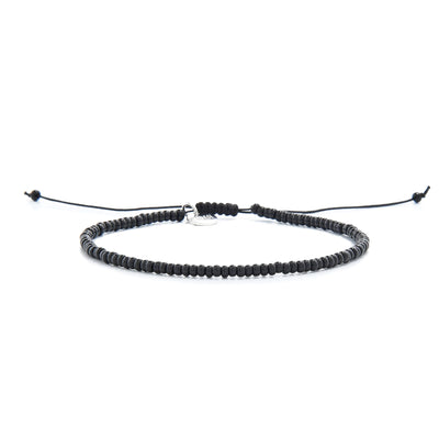 A full view of a simple black bead bracelet with an adjustable knot closure and a silver charm, lying on a white surface.
