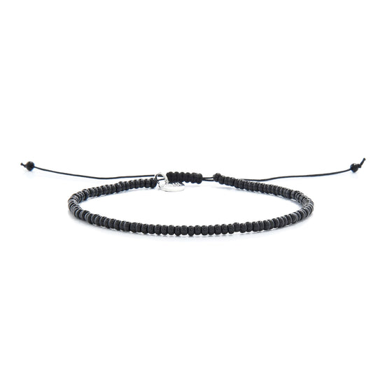 A full view of a simple black bead bracelet with an adjustable knot closure and a silver charm, lying on a white surface.