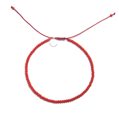 An angled view of an adjustable men beaded bracelet with red beads and a silver charm, lying on a white surface.