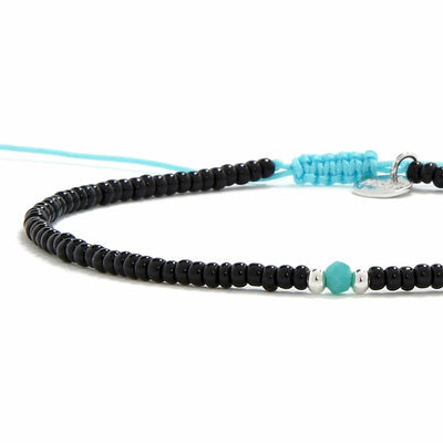 An angled view of a beaded bracelet featuring small black beads, a turquoise bead, a silver charm, and a bright blue adjustable cord.