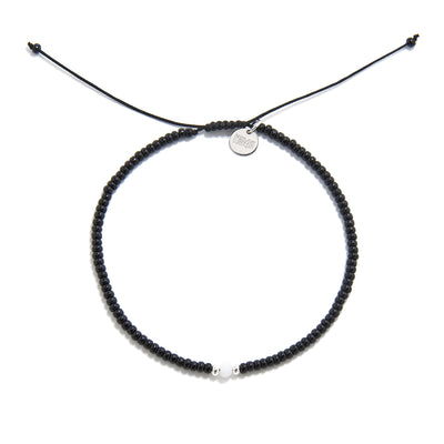 A top-down shot of a round bead bracelet black featuring a single white bead, a silver charm, and an adjustable cord.