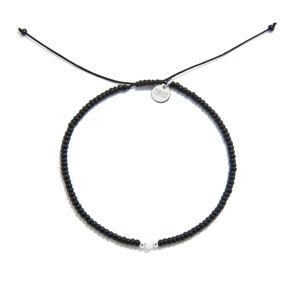 A top-down shot of a round bead bracelet black featuring a single white bead, a silver charm, and an adjustable cord.