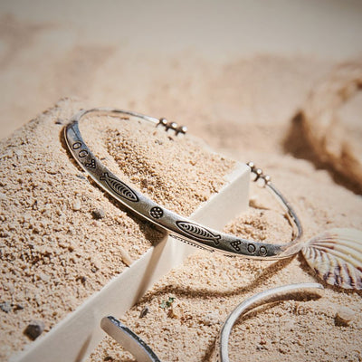Metal cuff bracelet with engraved fish pattern displayed on sand