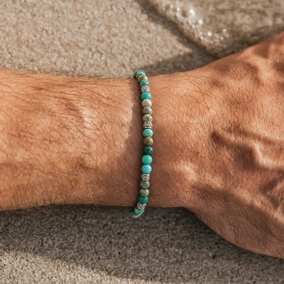 Green Jasper Bracelet worn on a wrist with turquoise beads, silver spacers, and green cord on a stone background.