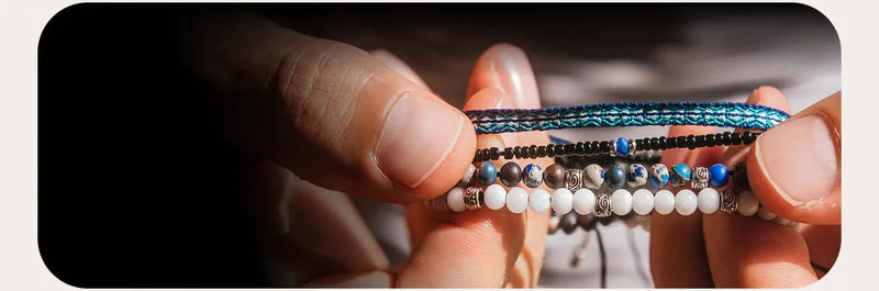 Handmade men’s bracelets held between fingers, showing stone beads, woven cord, and silver details.