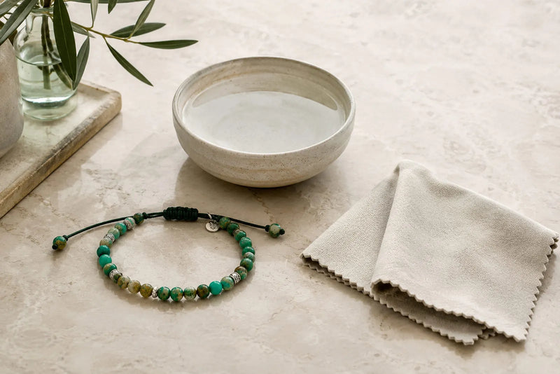 Green gemstone bead bracelet beside a soft cloth and bowl of water, showing a simple jewelry care setup on stone.