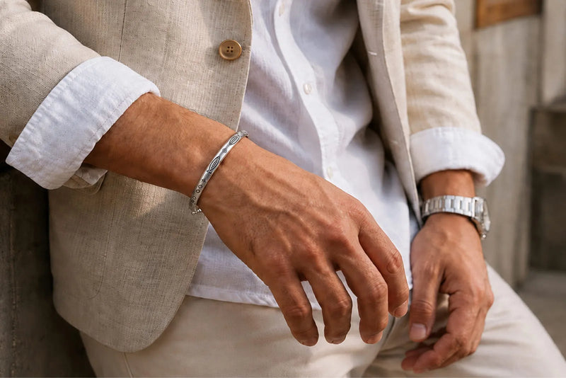 A man wears a silver cuff bracelet above the wrist bone with a beige linen suit and white shirt.