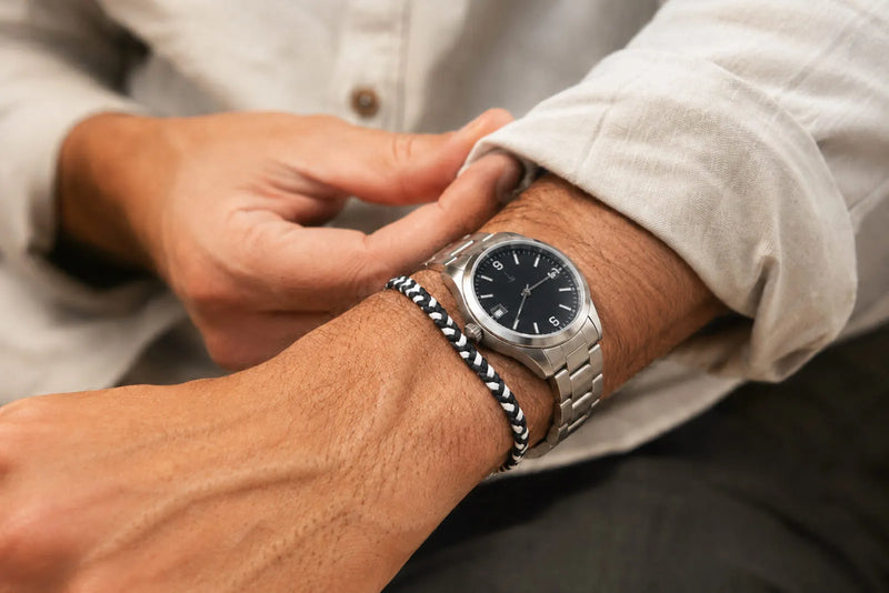 Man wearing a slim black and white woven bracelet beside a stainless steel watch with a neutral linen shirt.
