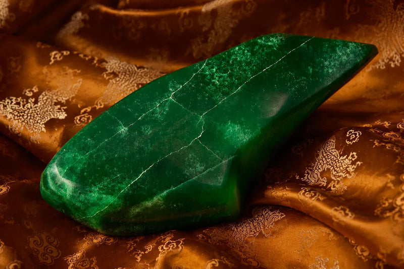 Jadeite vs Nephrite comparison featuring a polished deep green imperial jade stone displayed on golden patterned fabric.