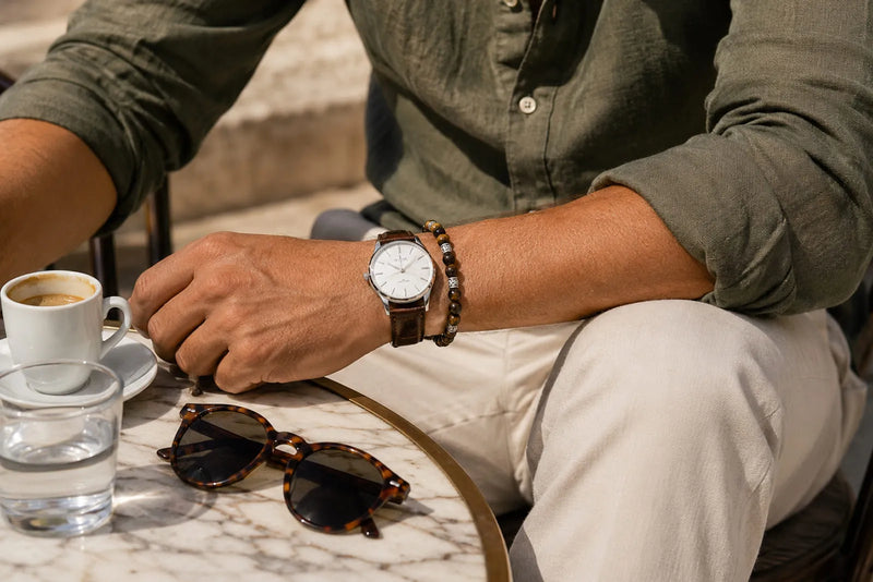Italian men’s fashion accessories with an olive shirt, cream trousers, leather watch, Tiger Eye bracelet, and sunglasses at a stone café table.