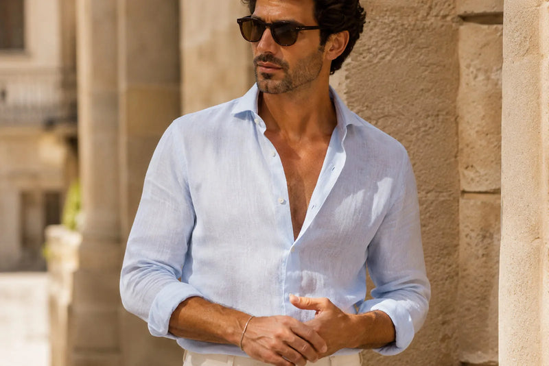 Italian men’s fashion close-up with a light blue linen shirt, rolled sleeves, sunglasses, and a slim bracelet in a warm Mediterranean setting.