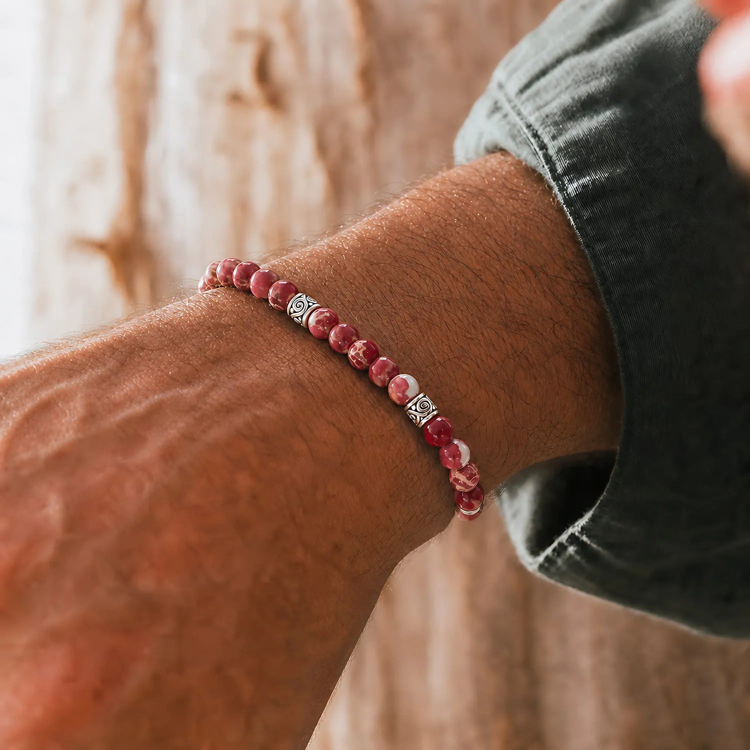 Red stone bracelet worn on a wrist with silver spacer beads and red cord against a warm wood background.