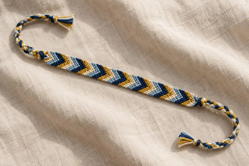 Navy, light blue, ivory, and yellow chevron bracelet resting on linen, with braided ties and knotted ends.