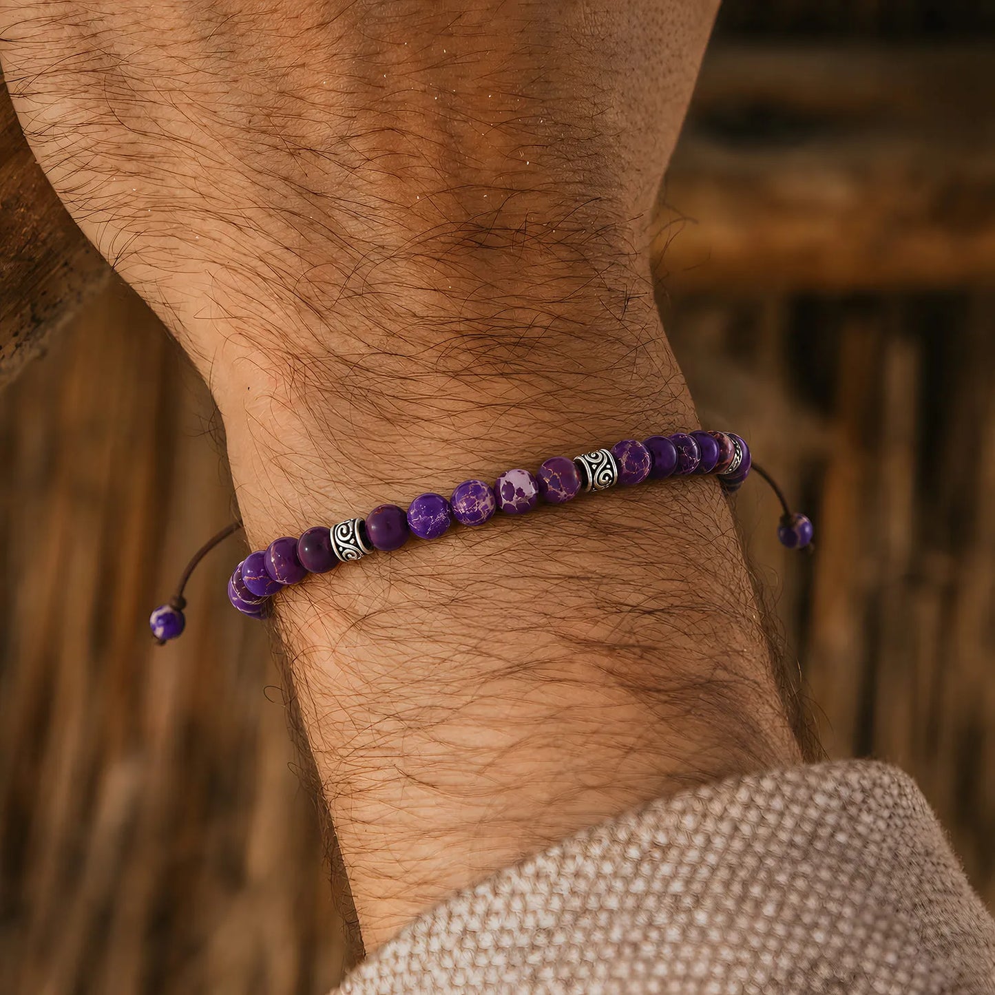Purple Jasper Bracelet on a wrist with silver spacer beads and brown cord against a warm wood background.
