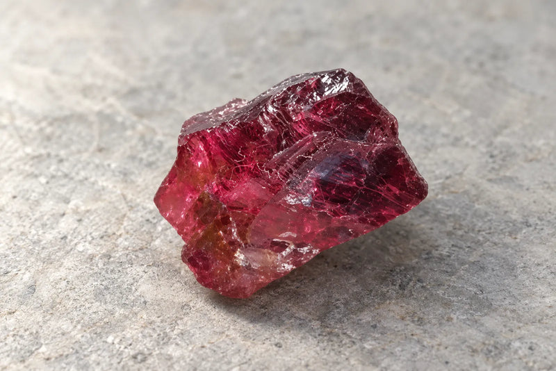 Rough raspberry-red rhodolite garnet with dark edges on a light stone surface.